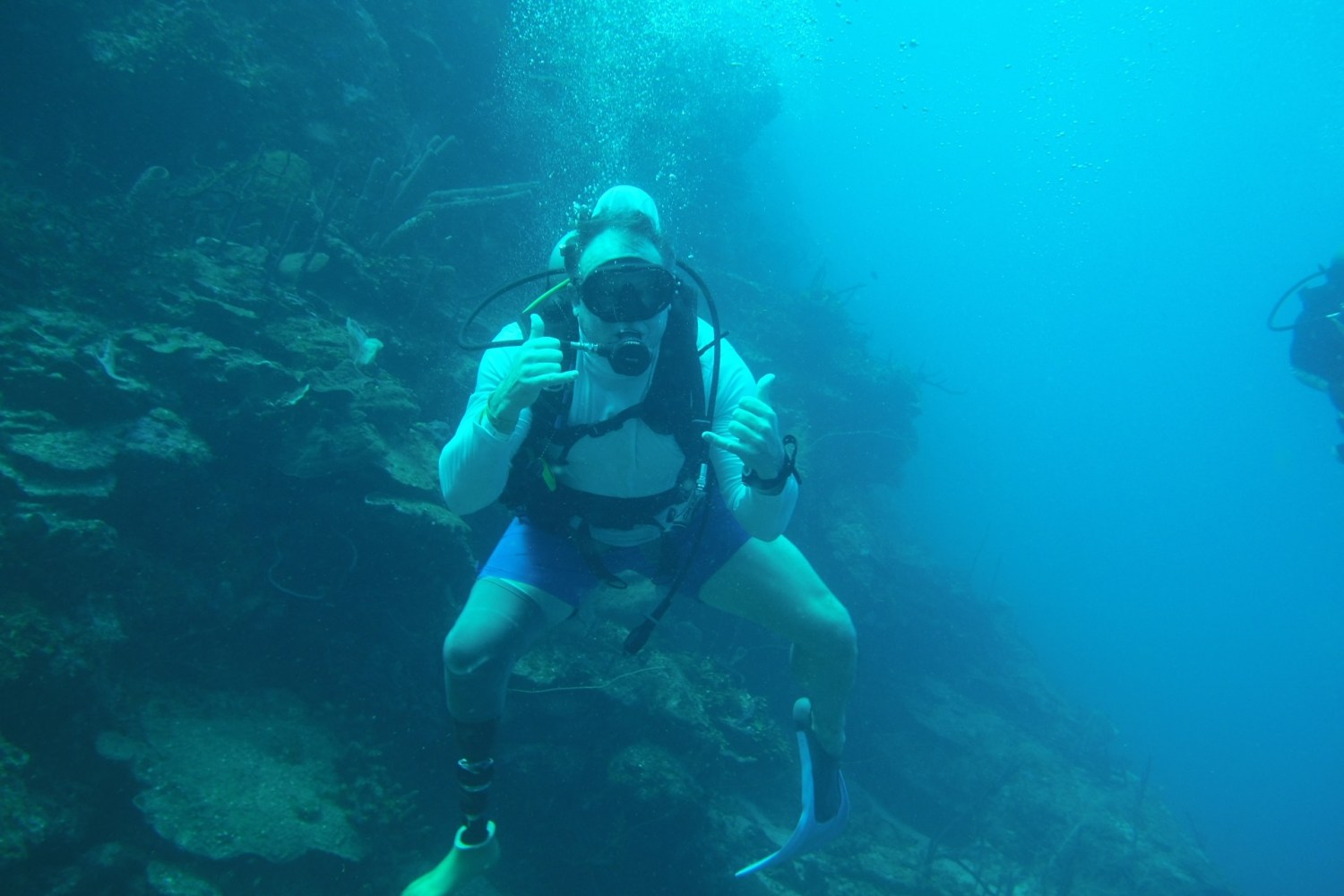 Diver underwater near a coral reef doing a hand gesture with scuba diving gear.