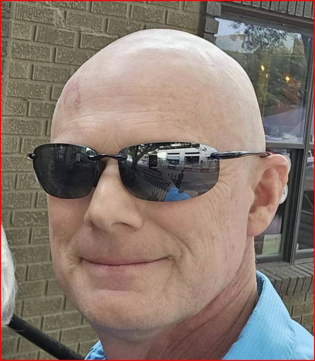 Bald person wearing sunglasses and a blue shirt, standing in front of a brick wall.