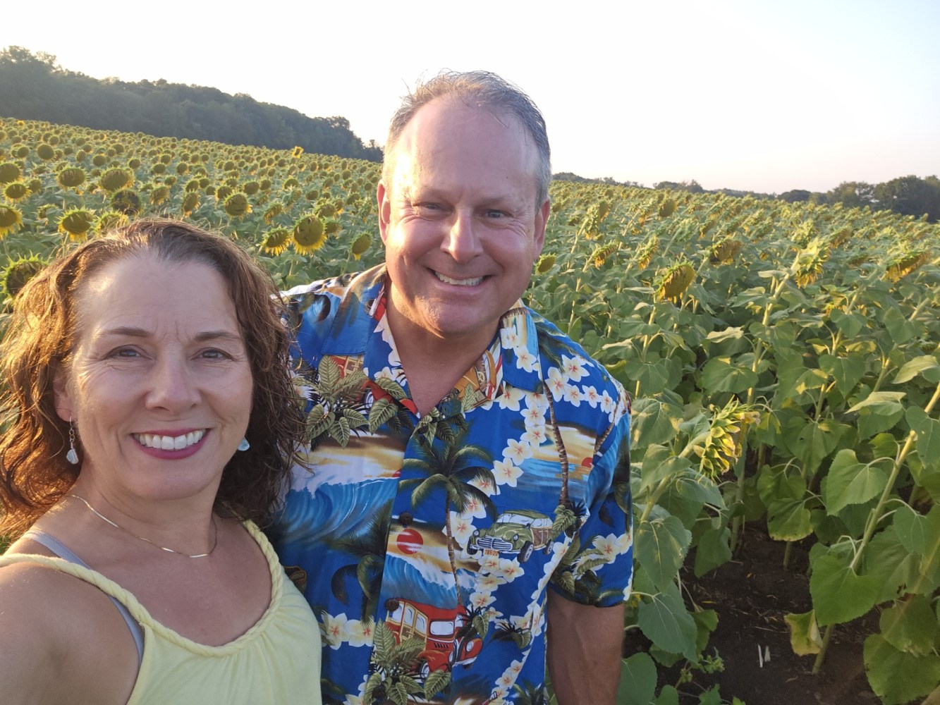 Smiling couple in front of a large sunflower field under a clear sky.