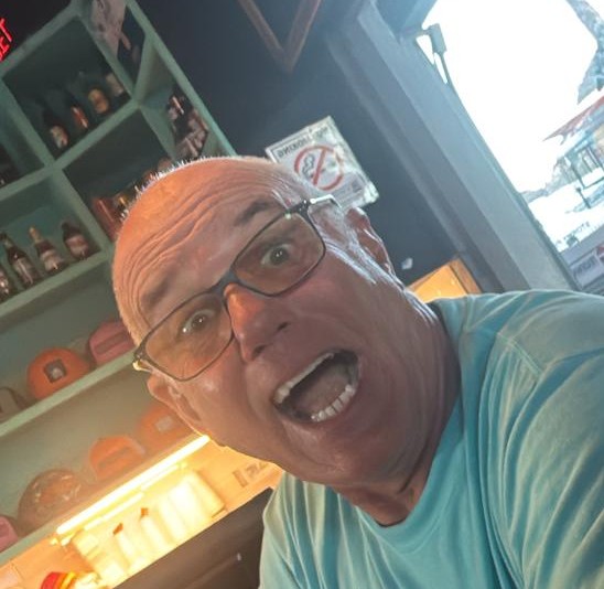 Man in glasses, open-mouthed, sitting in a bar with shelves and hats behind.