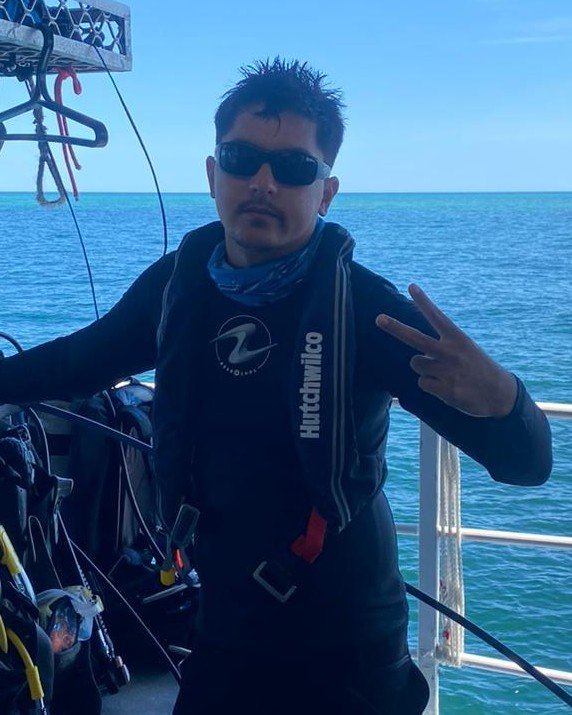 Man in scuba gear making a peace sign on a boat by the ocean.