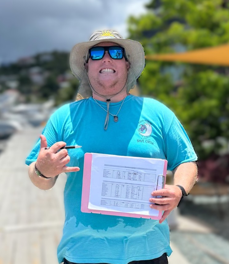Person in a hat and sunglasses holding a clipboard, wearing a blue T-shirt labeled 'DIVE CREW.'
