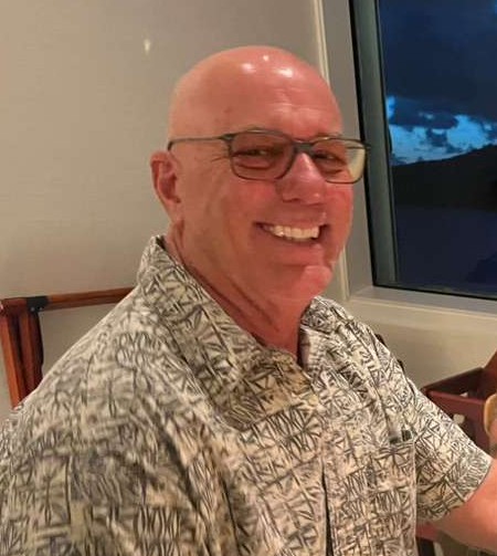 Smiling bald man with glasses in patterned shirt sitting indoors by a window.