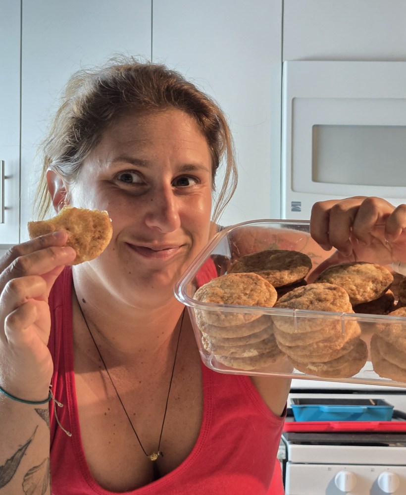 Person in pink tank top holding homemade cookies and smiling in a kitchen.